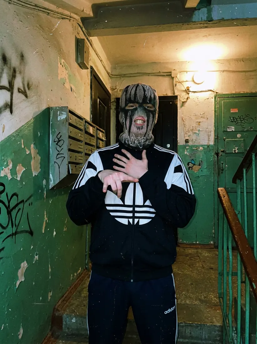 GLAKI in a balaclava and black Adidas track jacket in a green stairwell